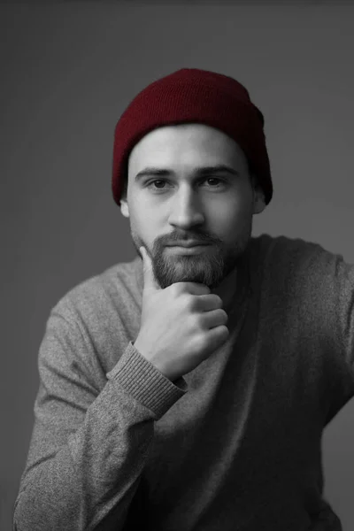 Portrait of a young bearded guy in a sweater, and a red hat, smiling. Studio photo on a gray background. Young handsome guy of twenty five years old, in a beige jacket and a burgundy hat, smiling, happy man, handsome bearded guy, actor, student, scho
