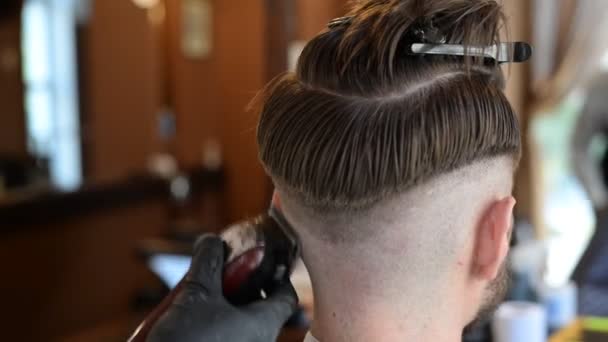 Seorang penata rambut memotong seorang pria muda berjanggut dengan pemangkas rambut, menyisir rambut di kepalanya. Karya master di mens potongan rambut di sebuah salon — Stok Video