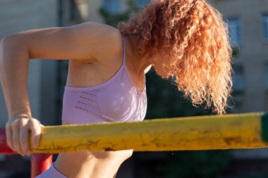Genç kızıl saçlı sporcu kadın sokaktaki düz olmayan barlarda şınav çekiyor. Pembe spor giyimli. Aktif yaşam tarzı, spor stili spor salonundaki düzgün olmayan barlarda şınav çeken kadın. Egzersiz yaşam tarzı kavramı