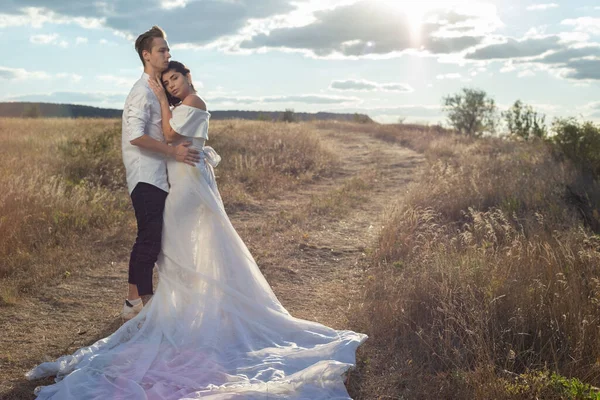結婚式のドレスで花嫁と新郎の若いカップル白いドレスは フィールドでは 曇りの青い空の背景に 空の背景を背景に 草原の中で結婚式の撮影 花嫁と新郎 — ストック写真