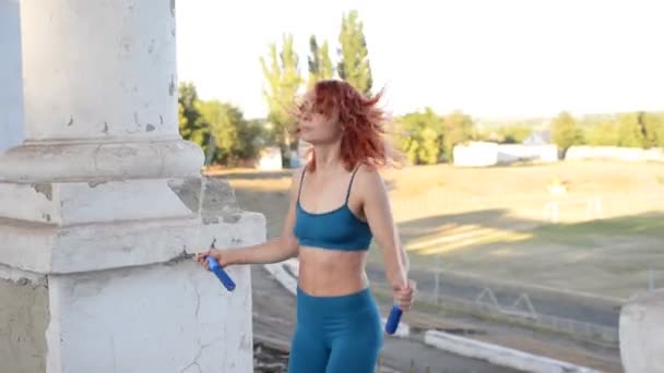 Young red-haired girl athlete jumping rope on the street. Active lifestyle. An athletic female body in leggings and a turquoise top, white sneakers, jumping in the air. — Stock Video