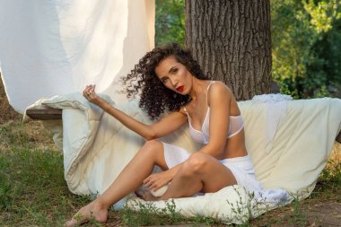 Genç, şehvetli, güzel bir esmerin portresi, beyaz iç çamaşırlı mutlu bir kız, gülümsüyor, lüks saçlarına dokunuyor, beyaz bir battaniyenin üzerinde oturuyor, yeşil çimlerin üzerinde, doğada. Çarşafları kurutmak için sapına asılı beyaz çarşafların arkasında. 