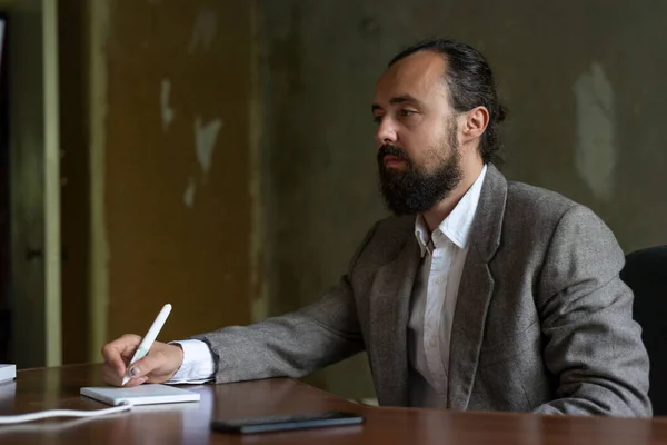 bearded young businessman in a business suit sitting at the computer table in the office. A computer mouse, writes a list in a graphical tablet. Office renovation Cheerful businessman sitting with laptop at office
