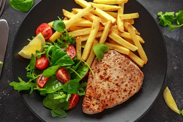 Stekt tonfisk biffar på svart tallrik med färsk grön, tomatsallad, citron och pommes frites. hälsosam mat — Stockfoto