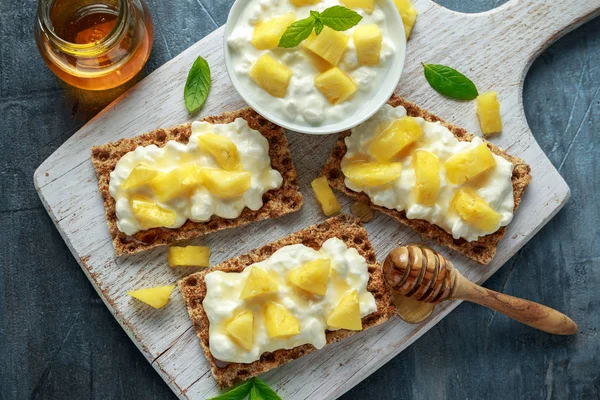 Tostadas caseras de pan crujiente con queso Cottage, piña y miel sobre tabla de madera blanca . —  Fotos de Stock