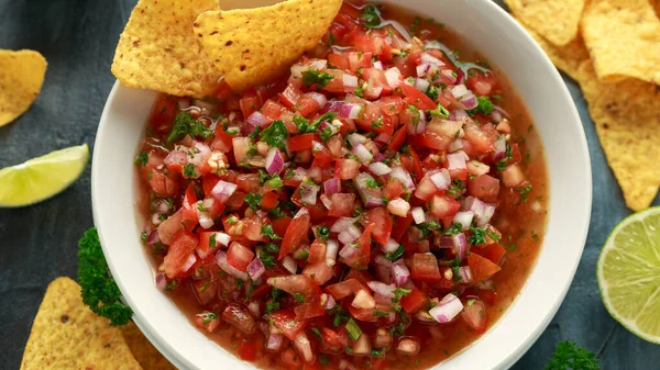 Mexican Tomato Salsa in white bowl with lime, red onion, jalapeno pepper, parsley and tortilla chips