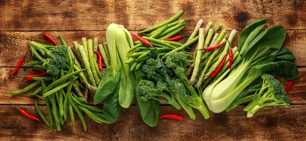 Fajta zöld zöldségek pak choi, spárga, brokkoli és madarak szem chili rusztikus fa asztalon — Stock Fotó