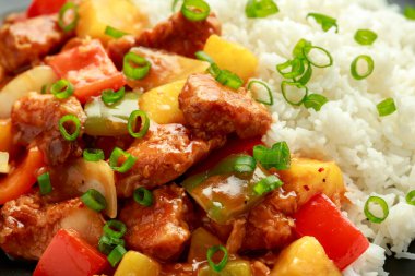 Tatlı ve ekşi soslu ananas ve tavuk yanında dolma biber, pirinç ve siyah tabakta taze soğan.