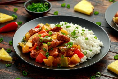 Tatlı ve ekşi soslu ananas ve tavuk yanında dolma biber, pirinç ve siyah tabakta taze soğan.