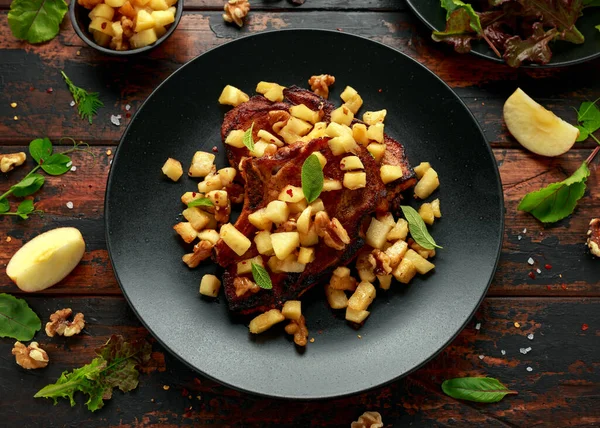 Chuletas de cerdo asadas con manzanas caramelizadas, nueces y salvia en plato negro sobre mesa de madera — Foto de Stock