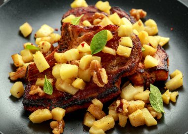 Kızarmış domuz pirzolası, karamelli elma, ceviz ve ahşap masada siyah tabakta adaçayı.