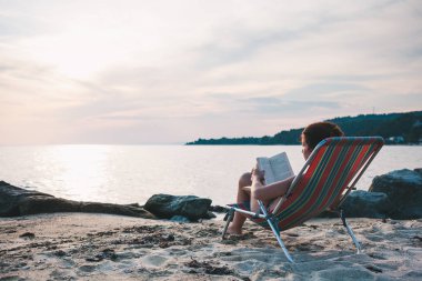 Genç bir kadın sahilde kitap okuyor.