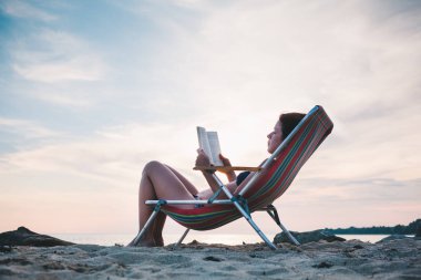 Genç bir kadın sahilde kitap okuyor.