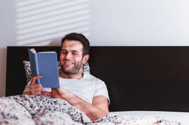 Genç adam yatakta kitap okuyor.