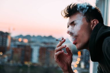 Young man smoking a cigarette on the balcony in sunset