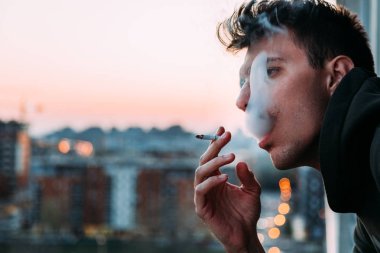 Young man smoking a cigarette on the balcony in sunset