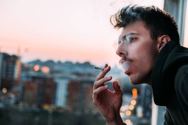 Young man smoking a cigarette on the balcony in sunset