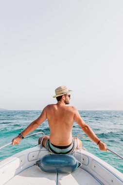 Teknenin pruvasunda oturan genç adam denizde manzaranın tadını çıkarıyor