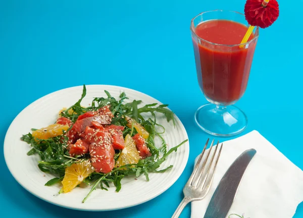 Kırmızı peçeteyle servis edilen roka salatası, portakal ve susamlı füme somon ve mavi arka planda domates suyu..