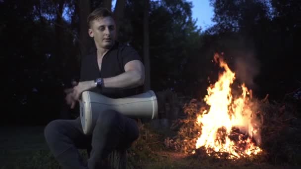 ハンサムな男が妖精の森の中で火の周りに座って太鼓を演奏する — ストック動画