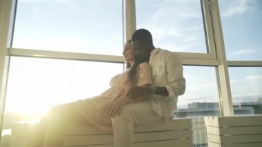 Yakışıklı siyah adam, güzel açık tenli kızı kucaklıyor pencere kenarında oturuyor, güzel bir manzarası var.