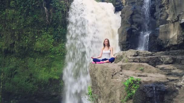 Femme assise dans la pose de lotus de yoga sur le bord à côté de la cascade Tegenungan — Video