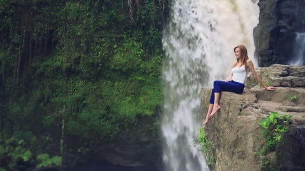 Woman sitting on a cliff next to Tegenungan Waterfall — Stock Video