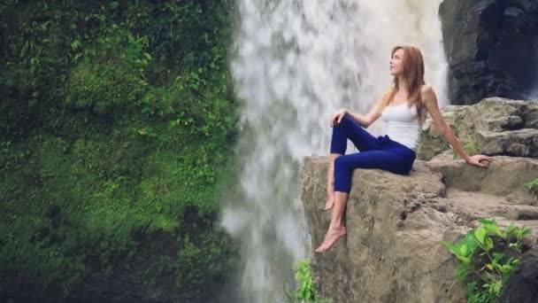 Frau sitzt auf einer Klippe neben dem Tegenungan-Wasserfall — Stockvideo