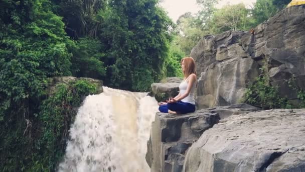 Kvinna som sitter i yoga lotus poserar på kanten bredvid Tegenungan Waterfall — Stockvideo