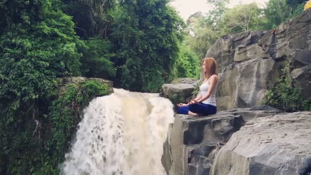 Donna seduta in posa loto yoga sul bordo accanto alla cascata Tegenungan — Video Stock