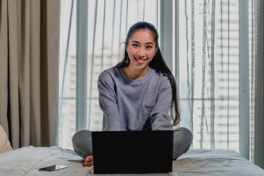 Açık mavi eşofmanlı Asyalı Çinli kız evde çalışıyor. Yatakta dizüstü bilgisayarla oturuyor ve kameraya gülümsüyor. İç mekan, ev.