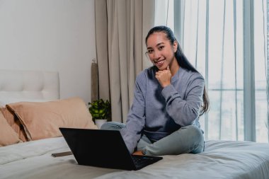 Açık mavi eşofmanlı Asyalı Çinli kız evde çalışıyor. Yatakta dizüstü bilgisayarla oturuyor ve kameraya gülümsüyor. İç mekan, ev.