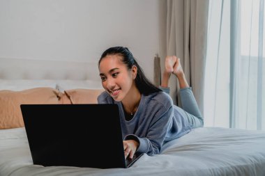 Açık mavi eşofmanlı Asyalı Çinli kız evde çalışıyor. Dizüstü bilgisayarla yatakta yatmak. İç mekan, ev.