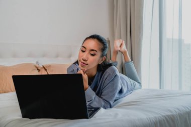 Asyalı Çinli açık mavi kazaklı bir kadın evde çalışıyor. Dizüstü bilgisayarla yatakta yatmak. İç mekan, ev.