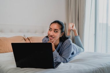 Açık mavi eşofmanlı Asyalı Çinli kız evde çalışıyor. Dizüstü bilgisayarla yatağa uzanıp düşünmek. İç mekan, ev.