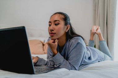 Açık mavi eşofmanlı Asyalı Çinli kız evde çalışıyor. Dizüstü bilgisayarla yatağa uzanıp düşünmek. İç mekan, ev.