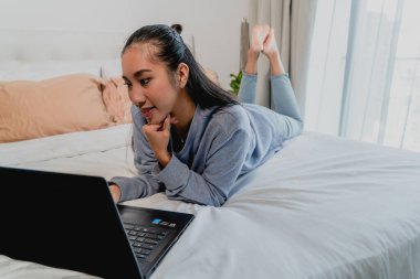 Asyalı Çinli açık mavi kazaklı bir kadın evde çalışıyor. Dizüstü bilgisayarla yatağa uzanıp düşünmek. İç mekan, ev.