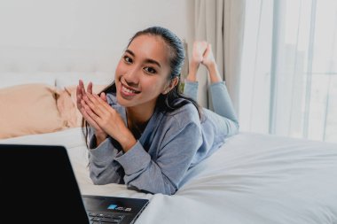 Açık mavi eşofmanlı Asyalı Çinli kız evde çalışıyor. Dizüstü bilgisayarla yatağa uzanıp düşünmek. İç mekan, ev.