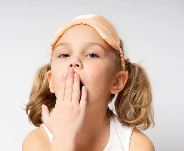 Ragazza allegra sbadiglia in pigiama in piedi su sfondo grigio in maschera del sonno al mattino . — Foto Stock