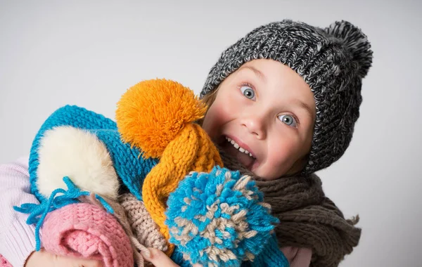 Little girl wearing knitted hat, scarf and sweater, holding a pile of hats, — Stock Photo, Image