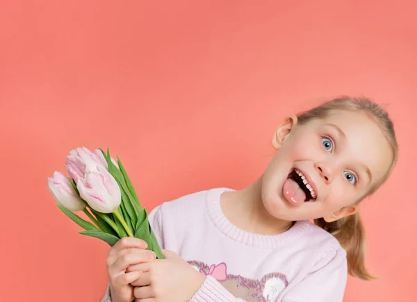 Pequena menina engraçada criança segura um buquê de tulipas como um presente para sua mãe e tolos ao redor na câmera, destaca sua língua. — Fotografia de Stock