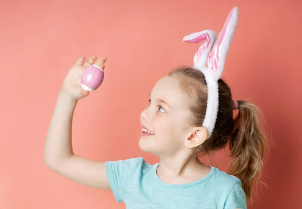 Little girl in funny bunny ears holds a multi-colored Easter egg, side view. — Stock Photo, Image