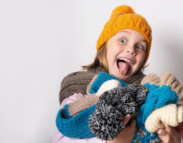 little girl wearing knitted hat, scarf and sweater, holding a pile of hats,