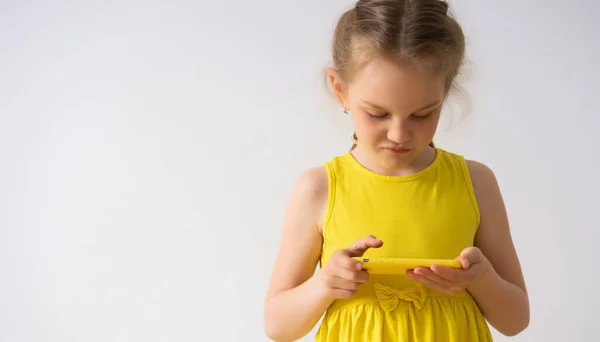 Atento niña rubia concentrada en vestido de verano amarillo se centra en la pantalla de su nuevo teléfono móvil. Primer plano estudio tiro aislado en blanco — Foto de Stock