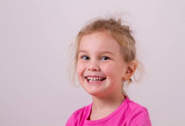 Close up shot of beautiful blonde Caucasian little girl wearing pink t-shit, looking at the camera with adorable smile, posing against white copy space concrete wall. Happy childhood concept