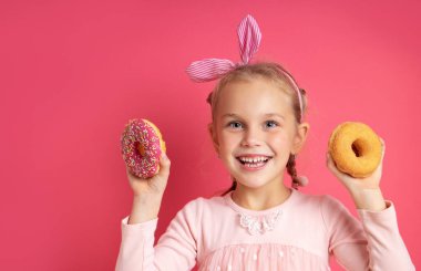 Resimli neşeli küçük kız pembe arka planda izole edilmiş renkli çöreklerle kamera önünde eğleniyor. Sevimli bir çocuğun gerçek olumlu duygularını ifade etmek. Metin için yer