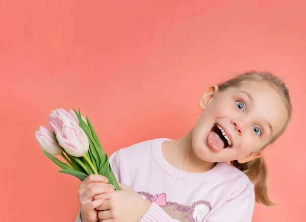 Pequena menina engraçada criança segura um buquê de tulipas como um presente para sua mãe e tolos ao redor na câmera, destaca sua língua. — Fotografia de Stock