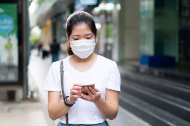 Asyalı genç bir kadın yüz maskesi takıyor, akıllı telefon kullanıyor ve Coperid-19 ya da Coronavirus salgını sırasında şehirde yürüyor. Sosyal uzaklık ve yeni normal yaşam tarzı kavramı