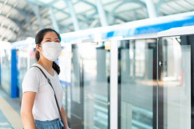 Yüz maskesi takan genç Asyalı bir kadın, akıllı telefon kullanıyor ve geçit istasyonunda duruyor, Coperid-19 ya da Coronavirus salgını sırasında gökyüzü üçlüsünü bekliyor. Sosyal uzaklık ve yeni normal kavram