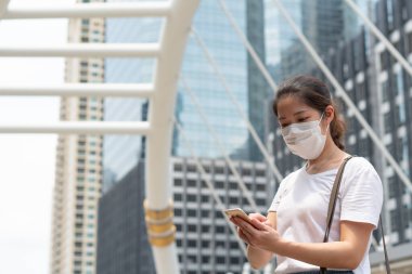 Asyalı genç bir kadın yüz maskesi takıyor, akıllı telefon kullanıyor ve Coperid-19 ya da Coronavirus salgını sırasında şehirde yürüyor. Sosyal uzaklık ve yeni normal yaşam tarzı kavramı